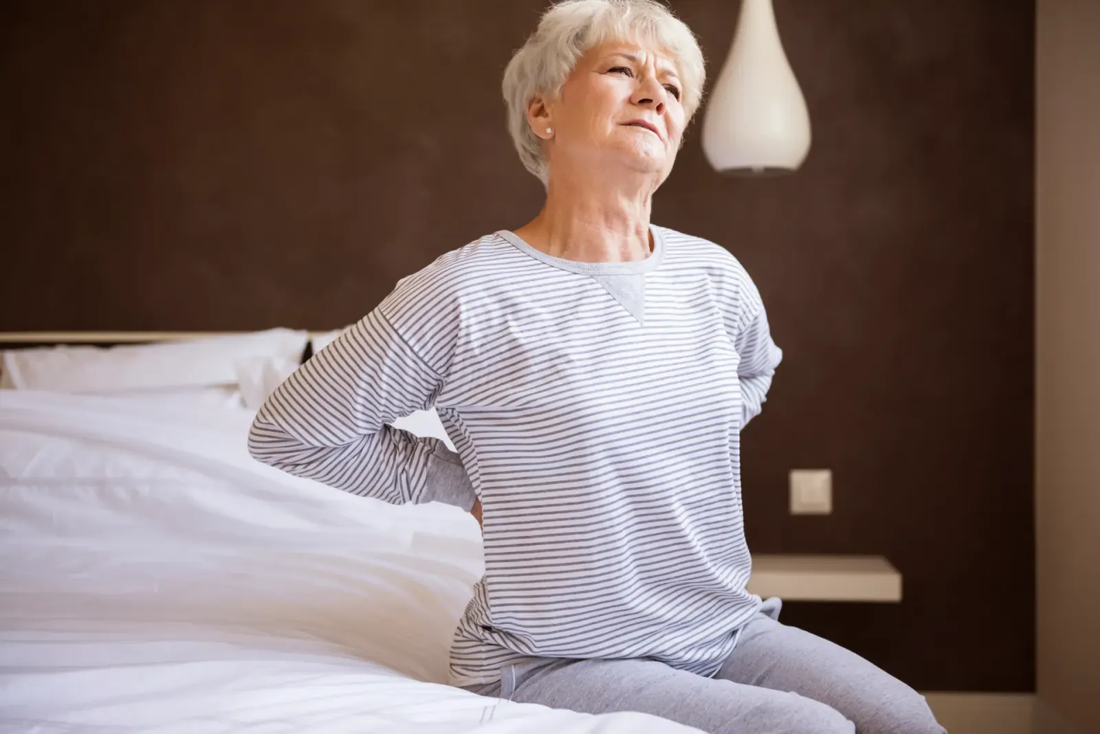 Older woman sitting on a bed holding her lower back