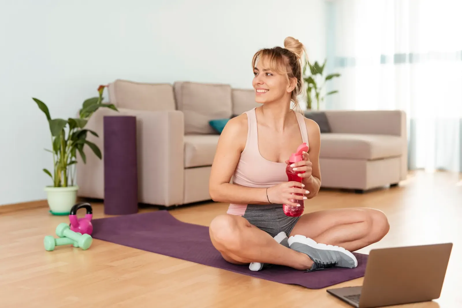 Woman sitting on a yoga mat at home with a water bottle and laptop
