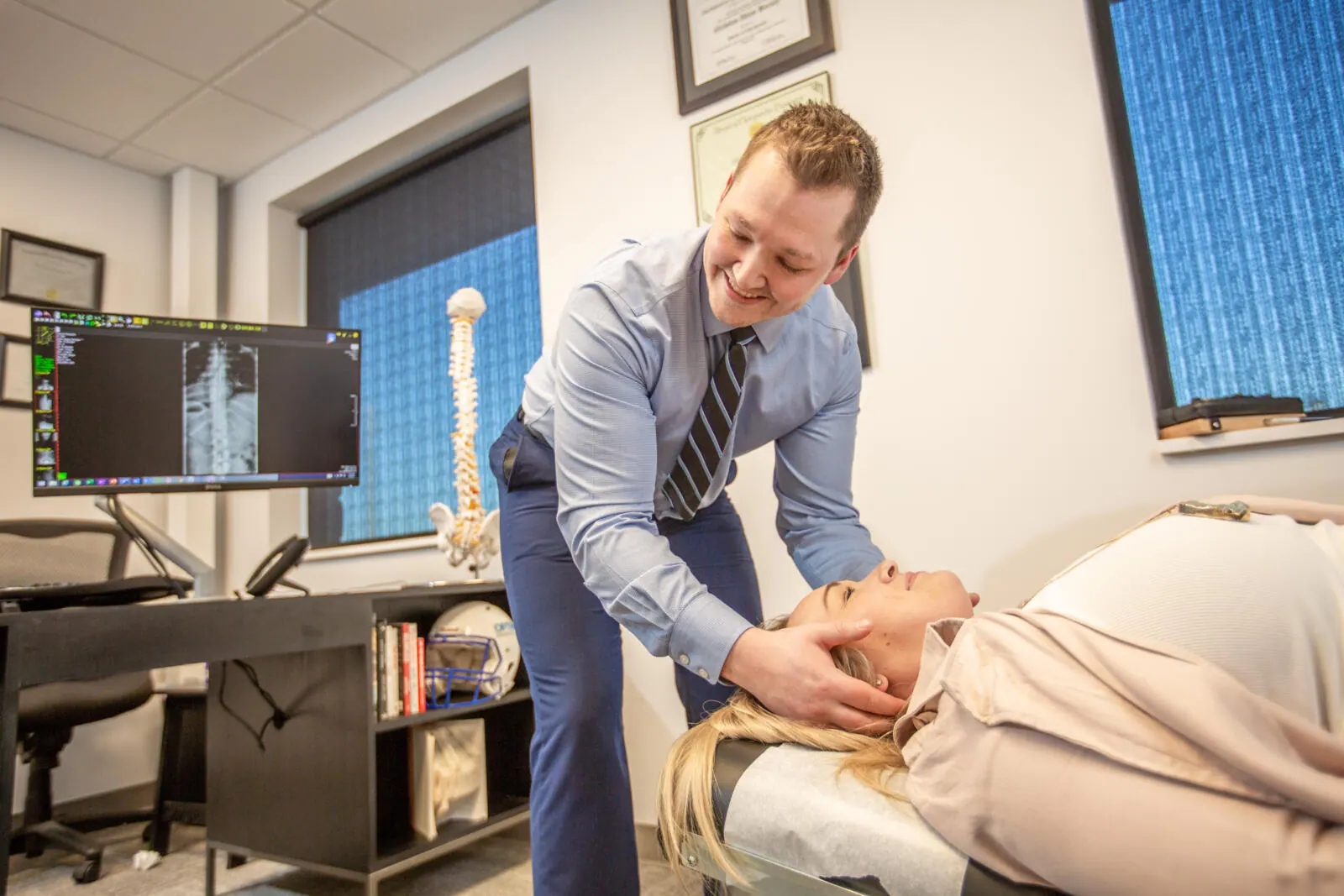 Chiropractor adjusting the neck of a female patient lying on a table, with a spine X-ray on the monitor behind