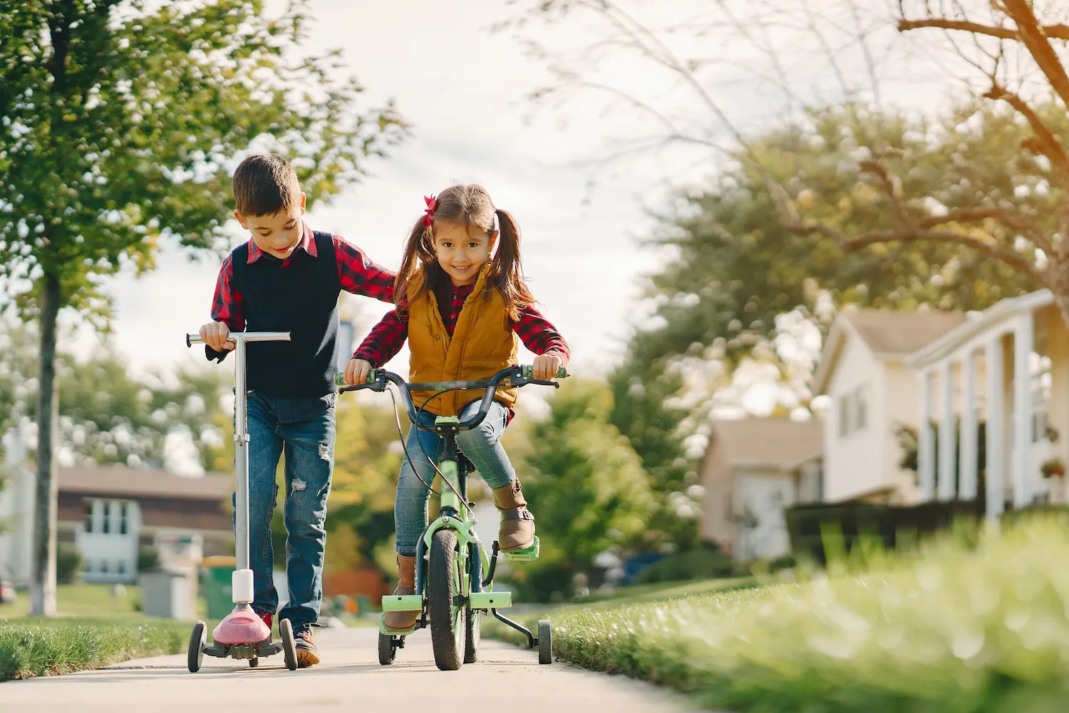 Boy on a scooter and girl on a bike riding along a neighborhood sidewalk