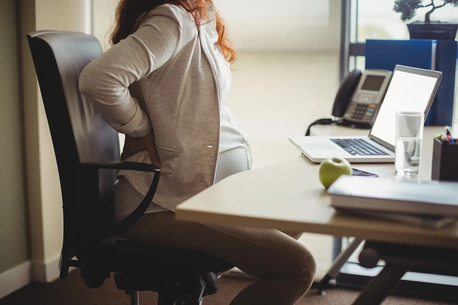 Pregnant woman at an office desk holding her lower back