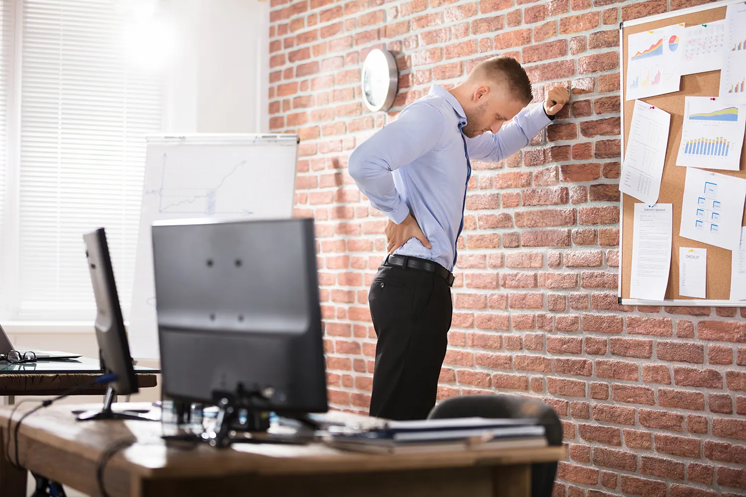 Man in an office leaning against a brick wall and holding his lower back